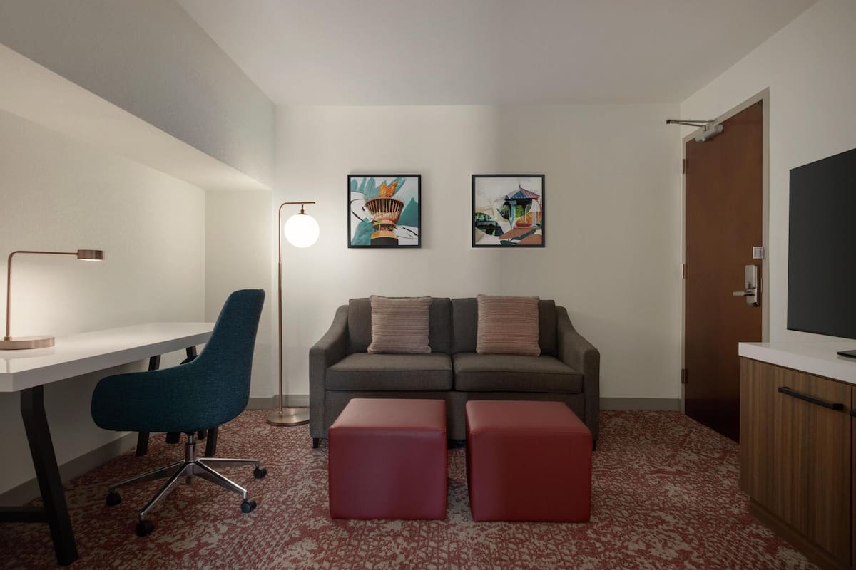 Una habitación con un escritorio, una silla de oficina azul, un sofá gris con dos otomanas rojas, una lámpara de pie y dos obras de arte enmarcadas en la pared.