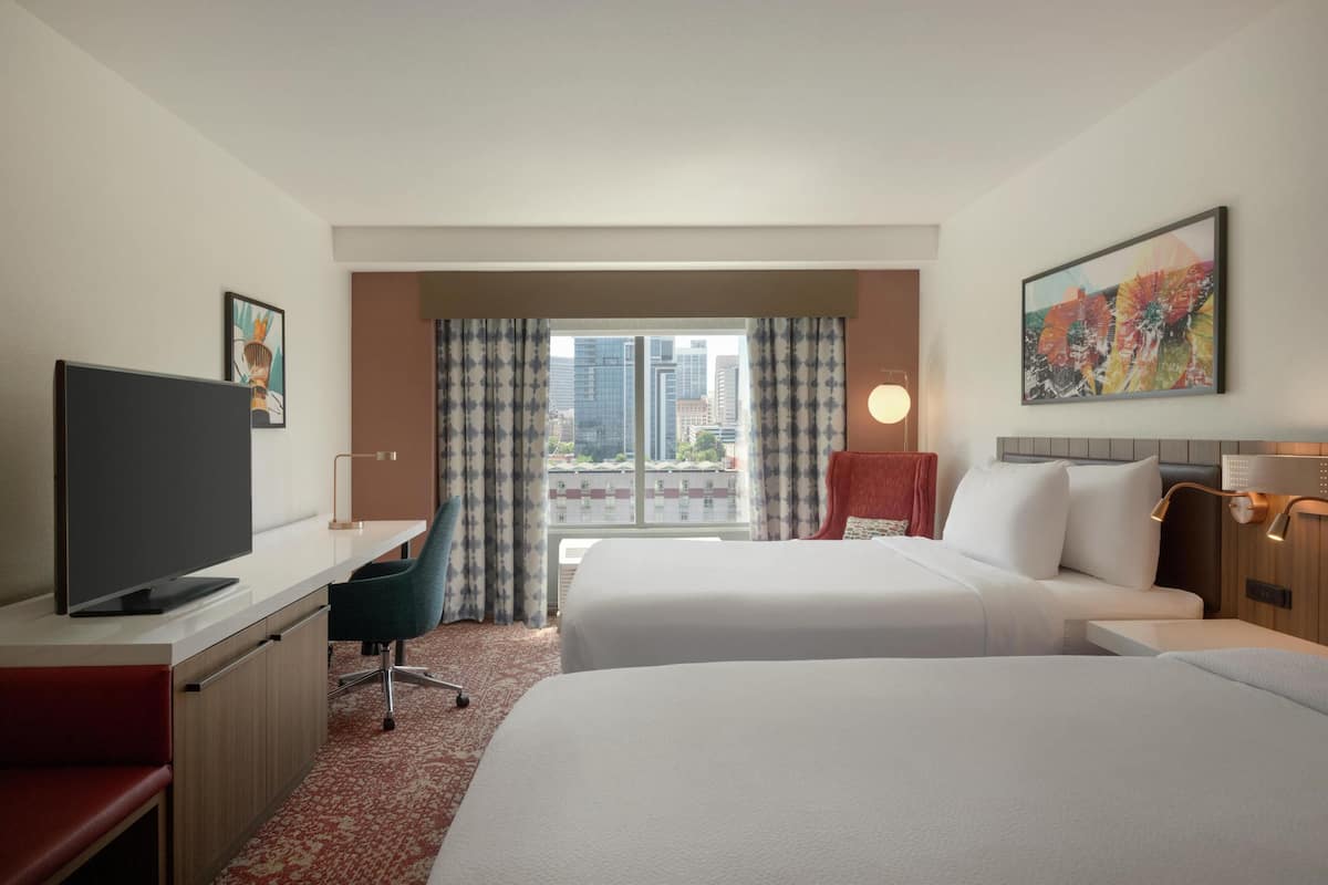 Habitación de hotel con dos camas, un escritorio con televisor, una silla, una ventana con cortinas y un cuadro en la pared.