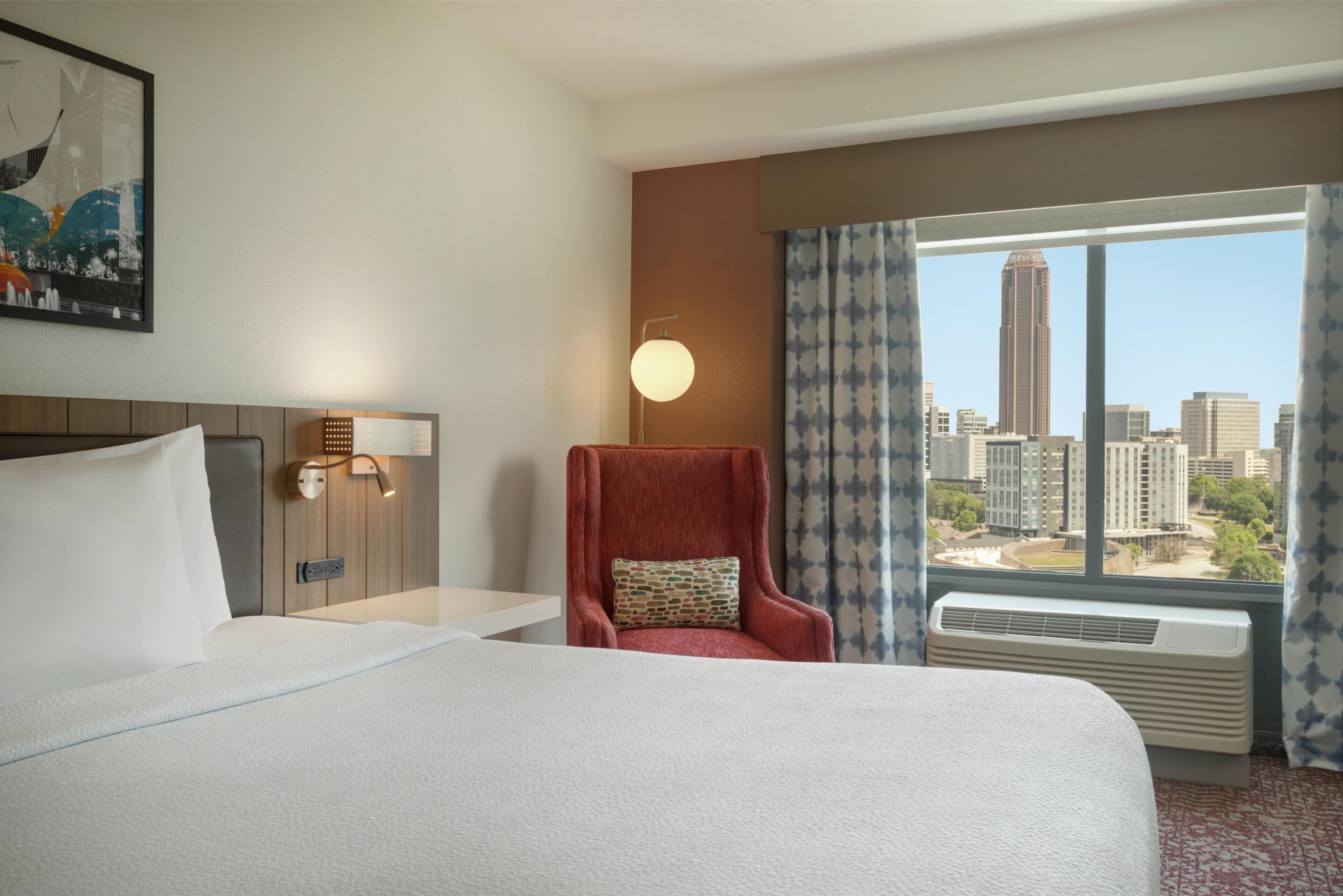 A hotel room with a large bed, a red armchair, a nightstand with a lamp, and a view of the city skyline through a window.