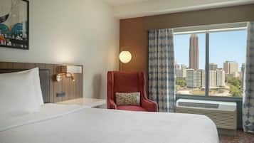 A hotel room with a large bed, a red armchair, a nightstand with a lamp, and a view of the city skyline through a window.