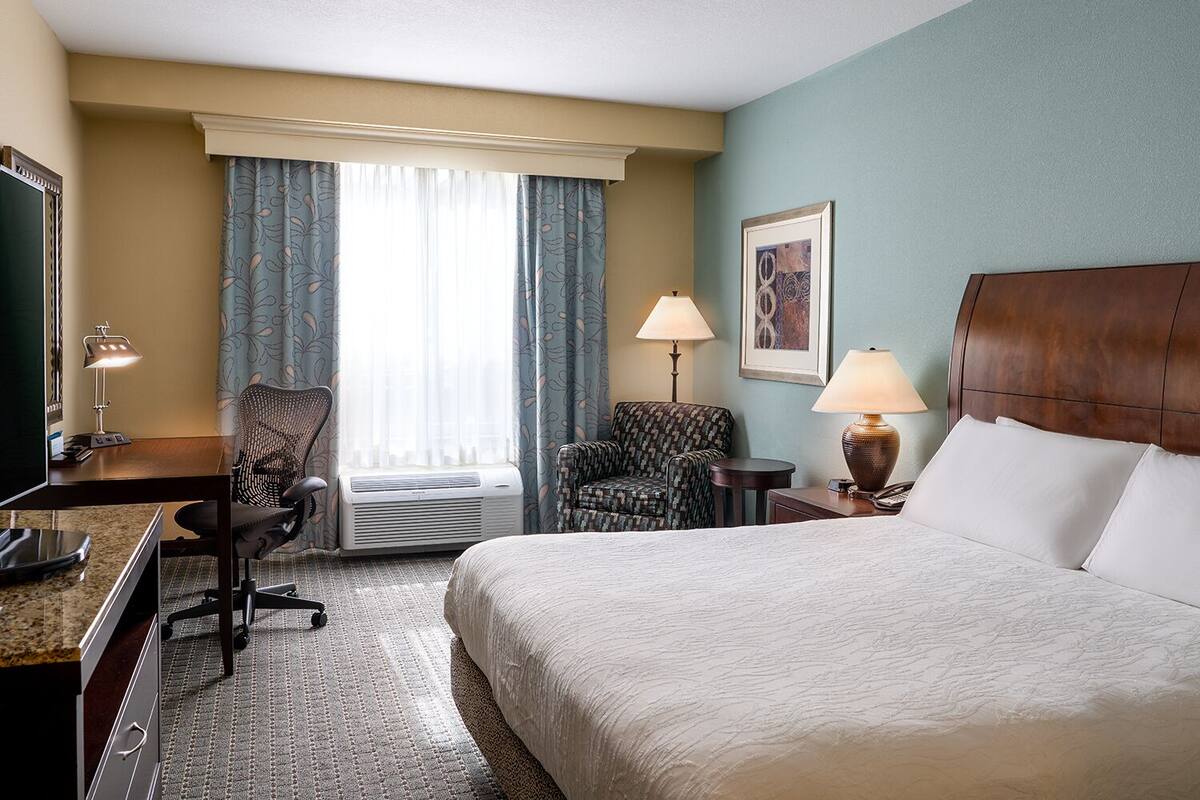 A hotel room with a bed, desk, chair, two lamps, and a window with curtains.