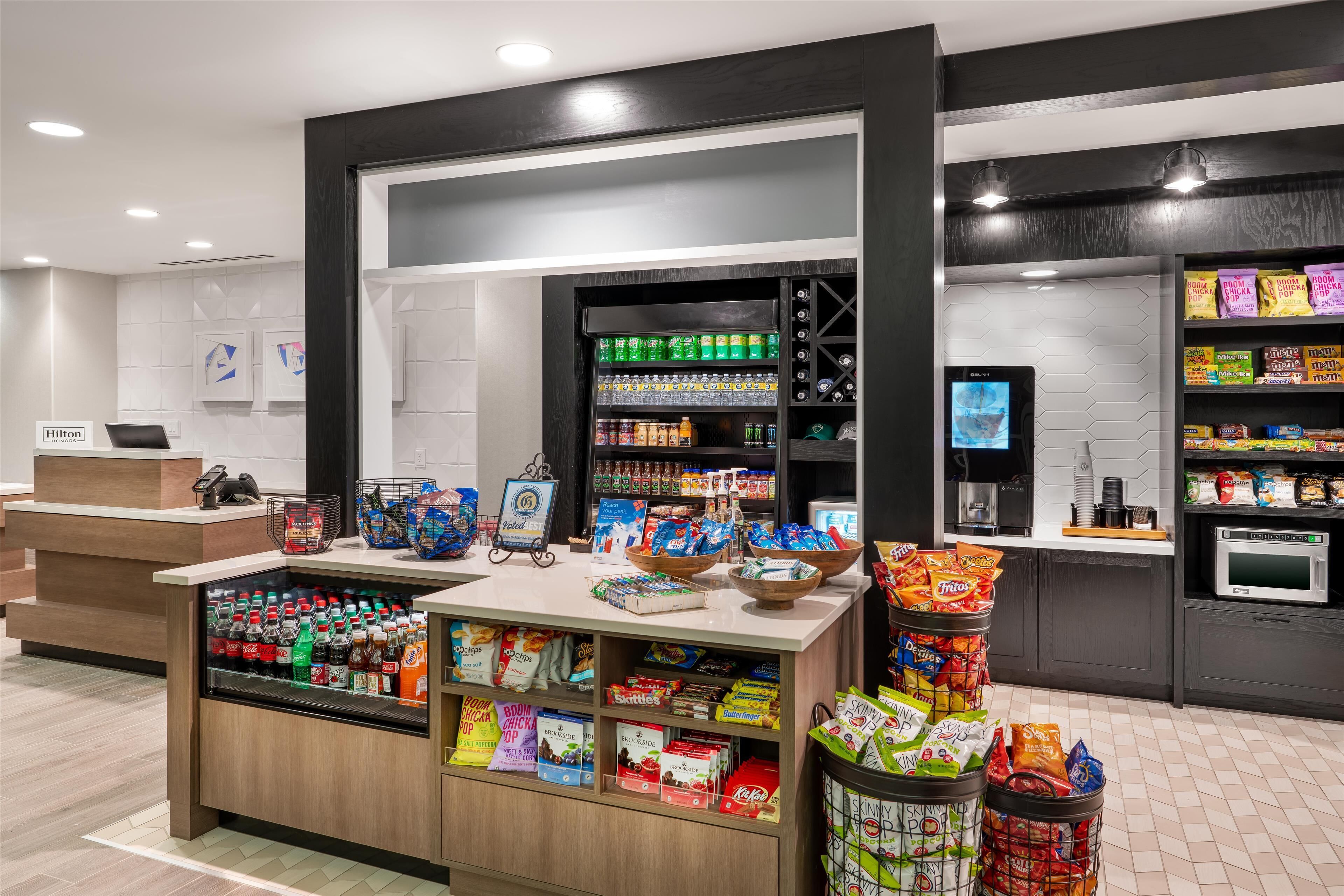 Un vestíbulo de hotel con una estación de bebidas y aperitivos autoservicio, surtida de diversas bebidas y bocadillos, una máquina de café y un microondas.