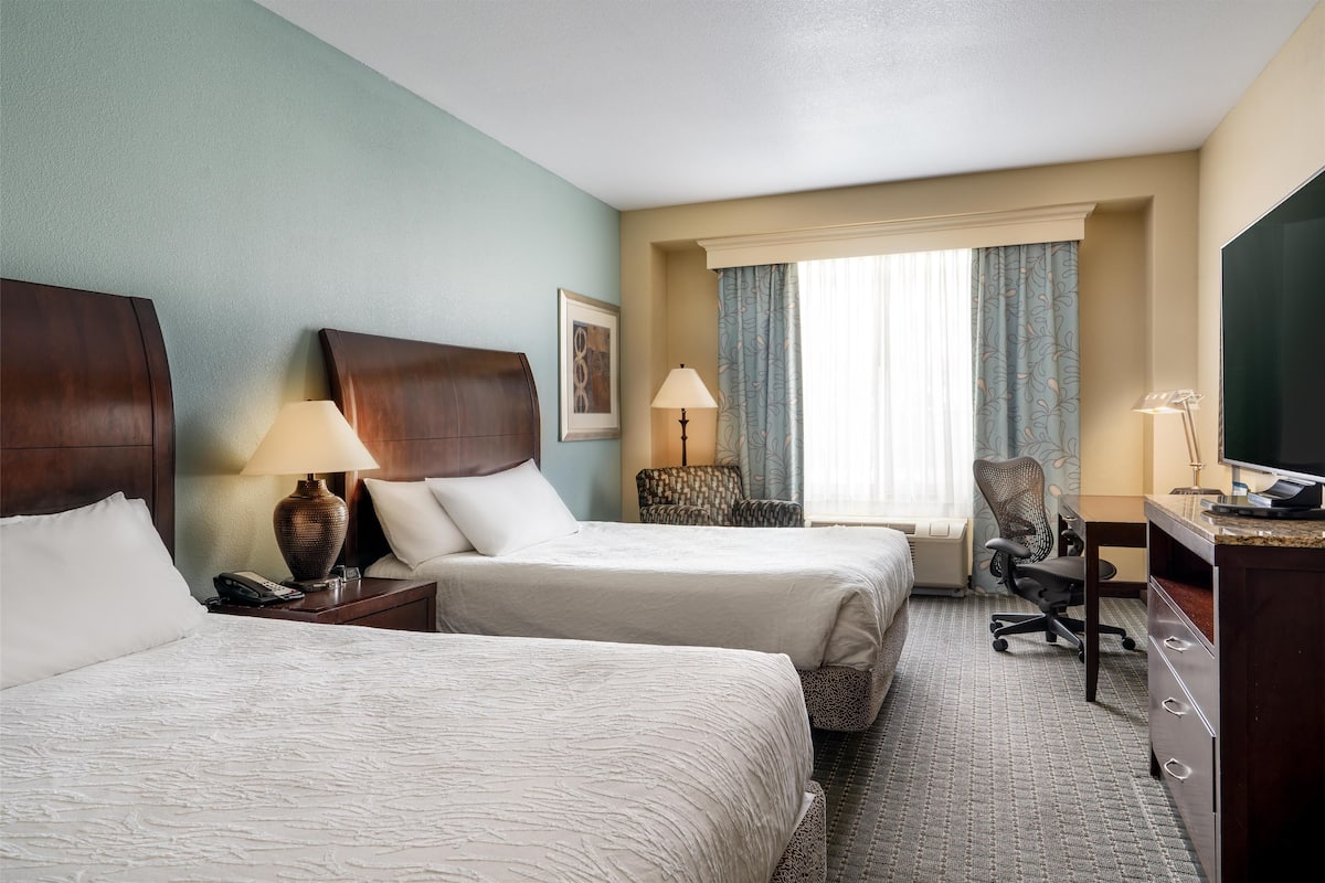 A hotel room with two beds, a desk, a chair, a TV, and a window with curtains.