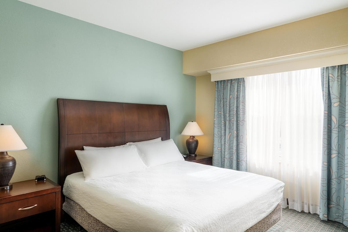 A hotel room with a large bed, two bedside tables with lamps, a window with curtains, and a headboard.