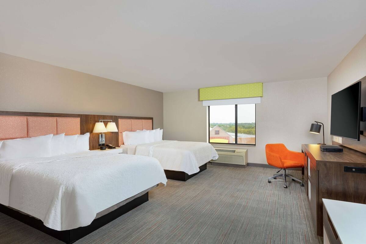 Habitación de hotel con dos camas, un escritorio, una silla naranja, un televisor y una ventana con vistas.