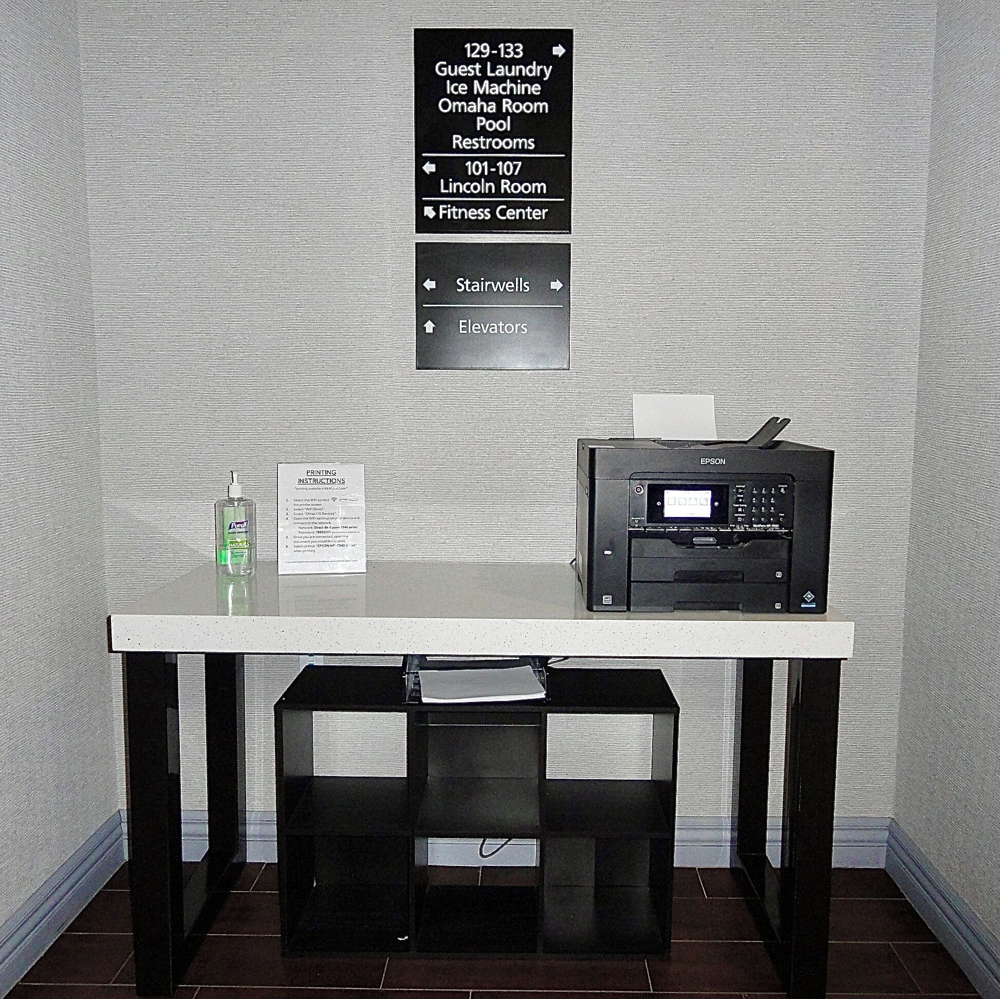 Quarto de hotel com uma mesa, uma impressora e um aviso indicando serviços e comodidades para hóspedes.