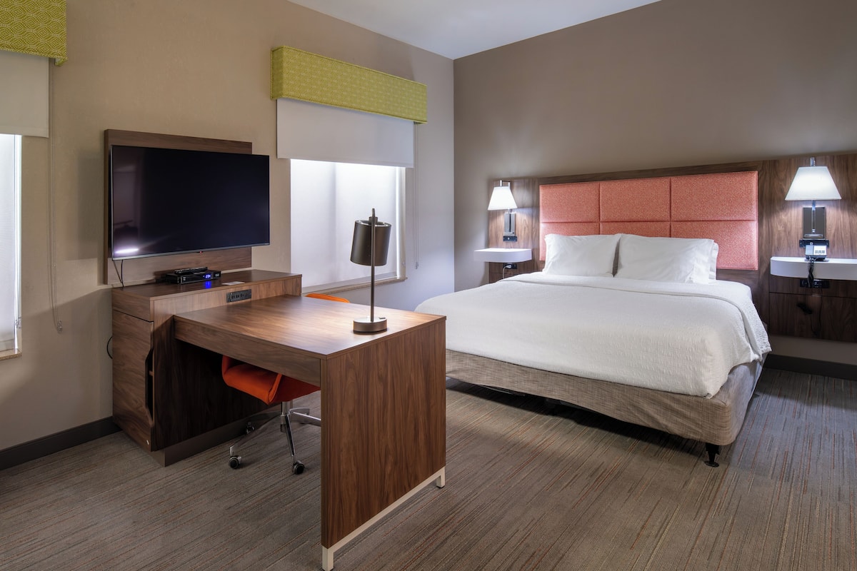 Habitación de hotel con una cama grande, un escritorio con televisor de pantalla plana, una lámpara y una ventana con persianas.