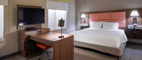 A hotel room with a large bed, a desk with a flat-screen TV, a lamp, and a window with blinds.