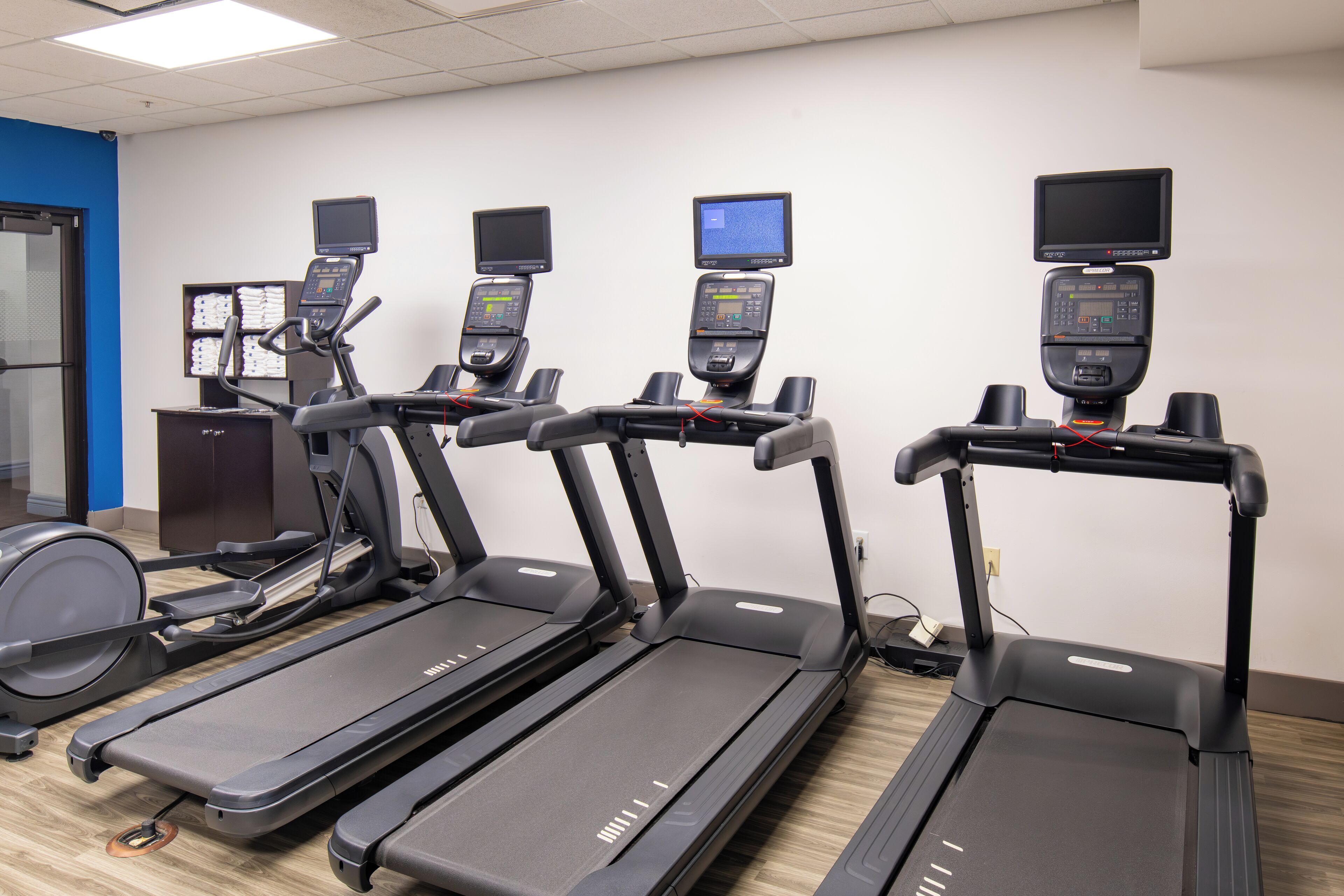 Four treadmills with attached screens in a room with a blue wall and wooden flooring.