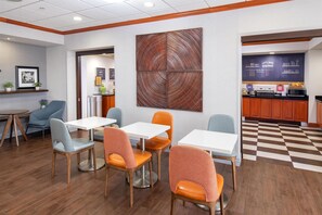 A dining area with wooden flooring, white tables, and orange chairs. There is a kitchen area with a checkered floor and a chalkboard with writing.