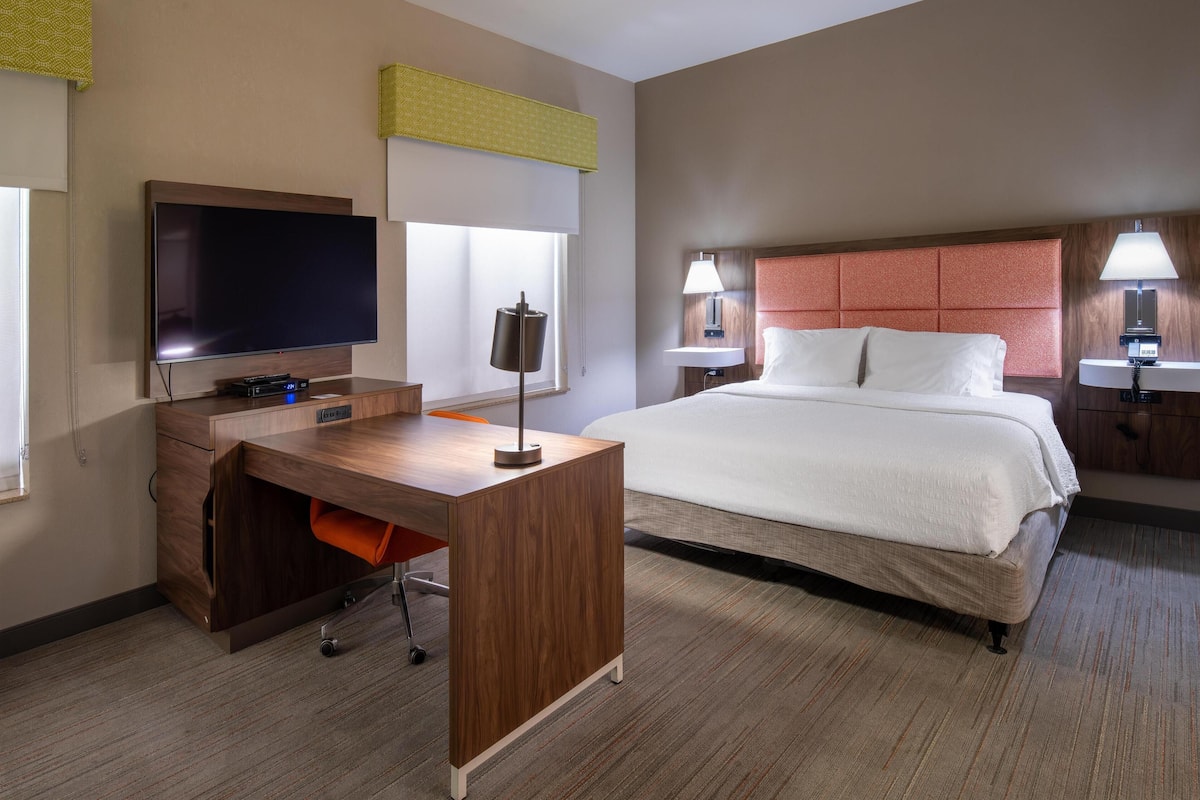Una habitación de hotel con una cama, un escritorio con televisor, una lámpara y una ventana.