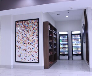 A hallway with a large geometric art piece on the left, wooden shelving units, and two refrigerated display cases with various products. - Holiday Inn Express & Suites Mansfield by IHG (Mansfield)