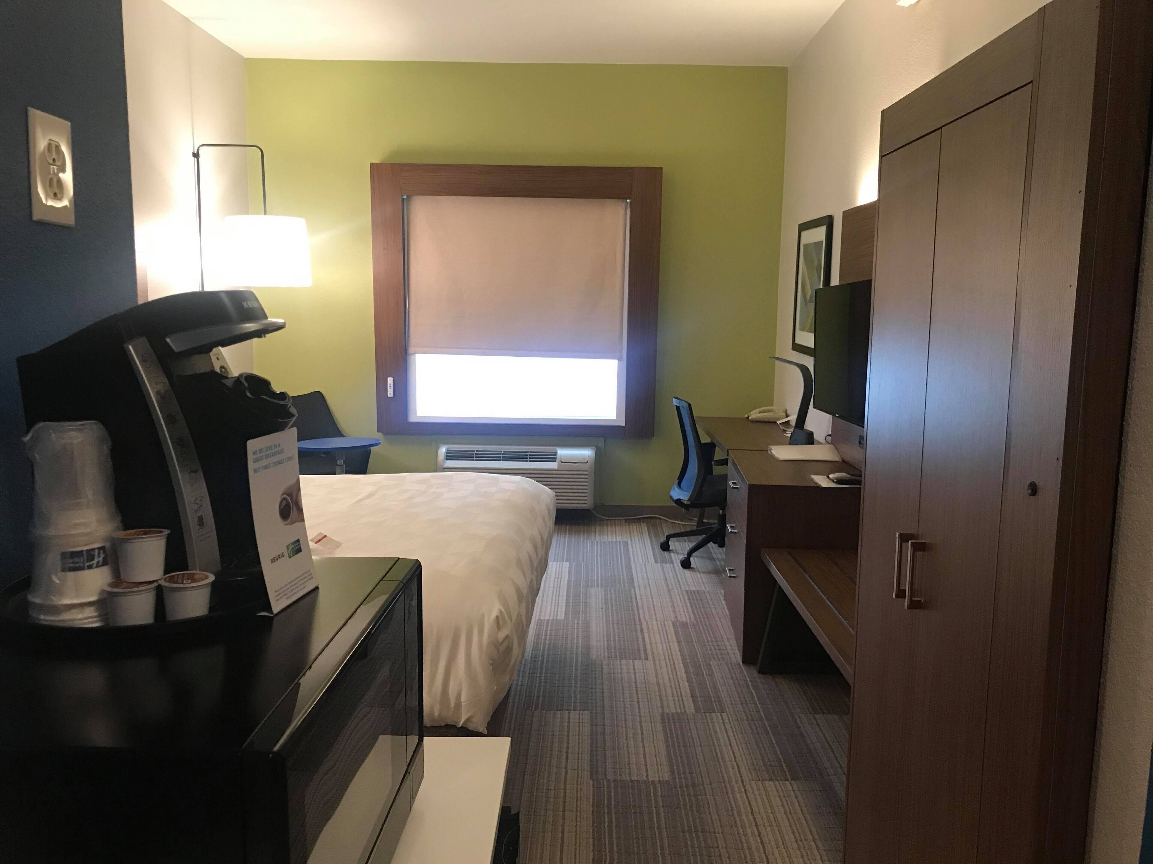 Ein Hotelzimmer mit Bett, Schreibtisch, Stuhl und Kleiderschrank. Ein Fenster mit Jalousien, ein Heizgerät und eine Lampe sind vorhanden.