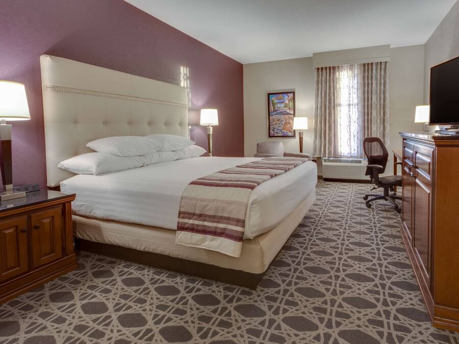 A hotel room with a large bed, two bedside tables with lamps, a desk with a flat-screen TV, and a patterned carpet.