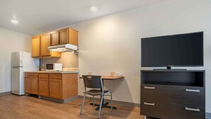 A compact kitchen with wooden cabinets, a microwave, a refrigerator, a chair, and a TV on a stand.