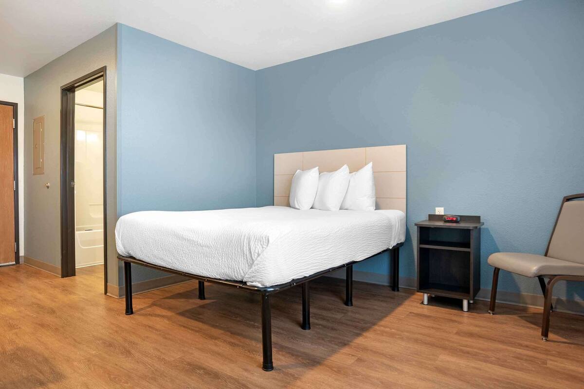 Habitación de hotel ordenada con cama, una silla, una mesita de noche y una puerta.