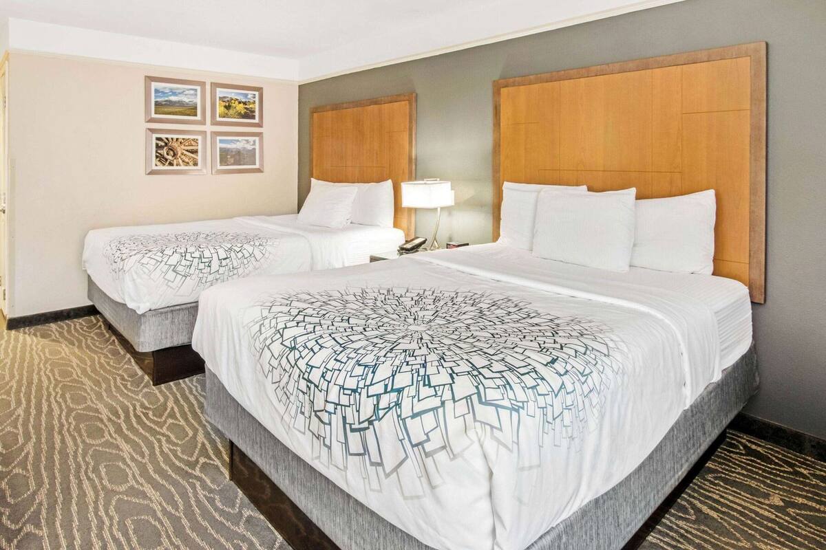Quarto de hotel com duas camas, uma cabeceira de madeira, carpete estampado e quadros nas paredes.