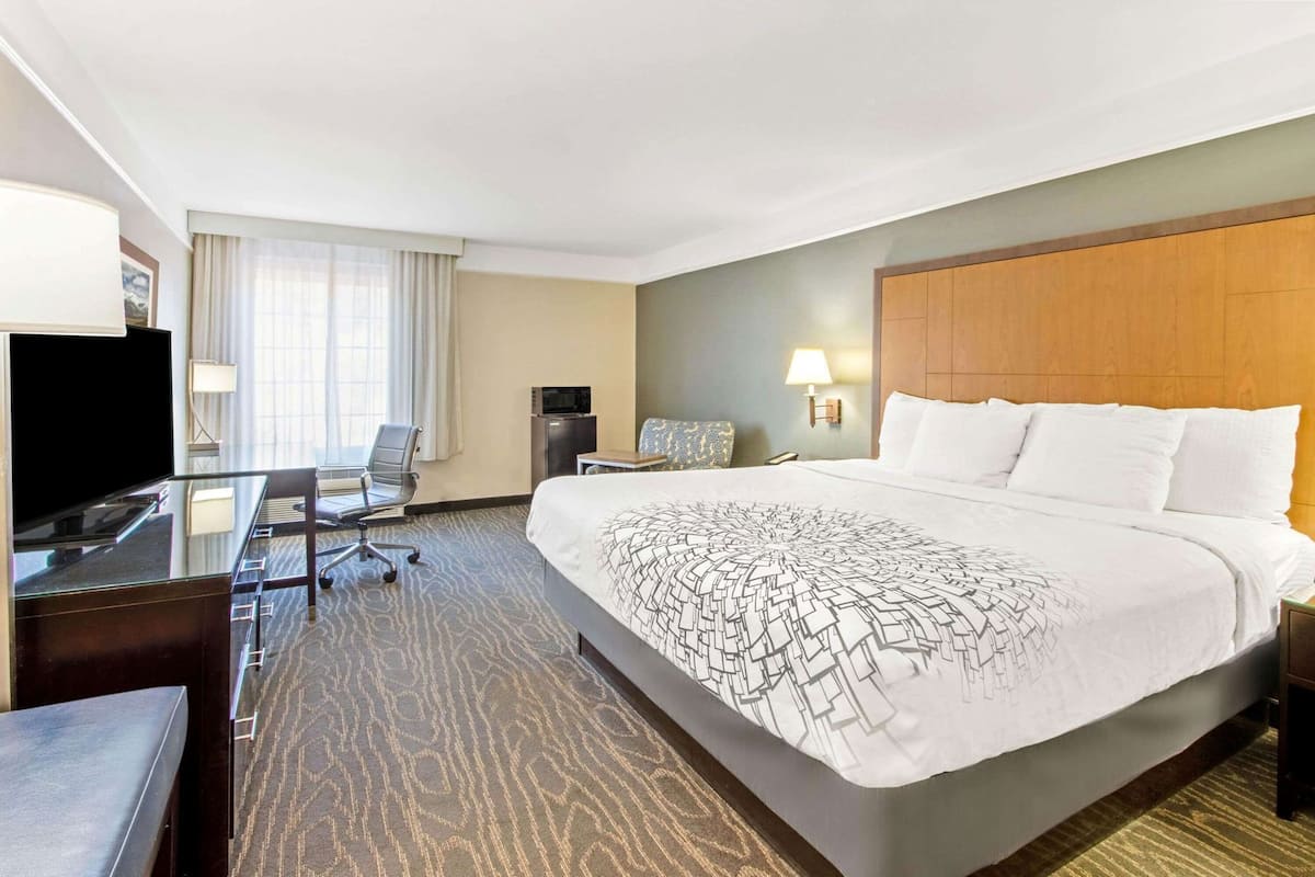 Quarto de hotel com uma cama grande, uma escrivaninha com cadeira, uma televisão e uma luminária.