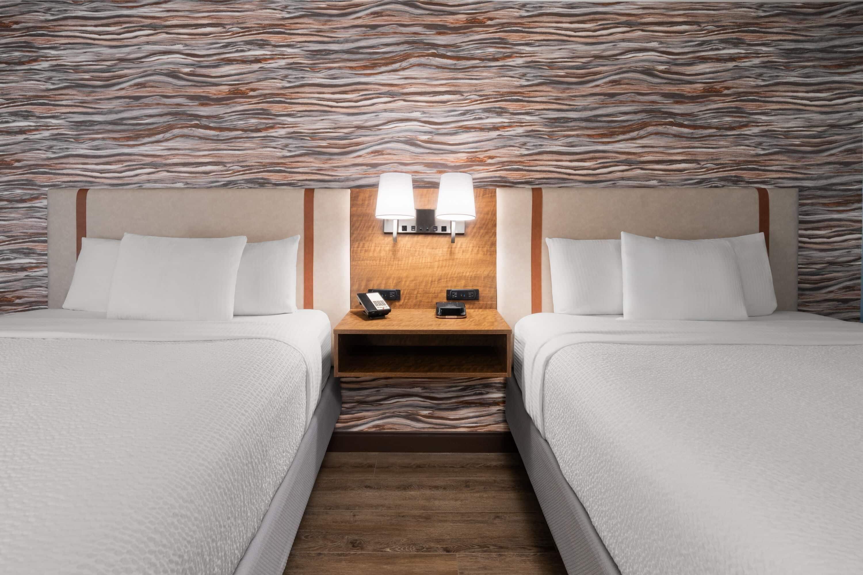A hotel room with two beds, a wooden nightstand with a phone and a lamp, and a textured wall.