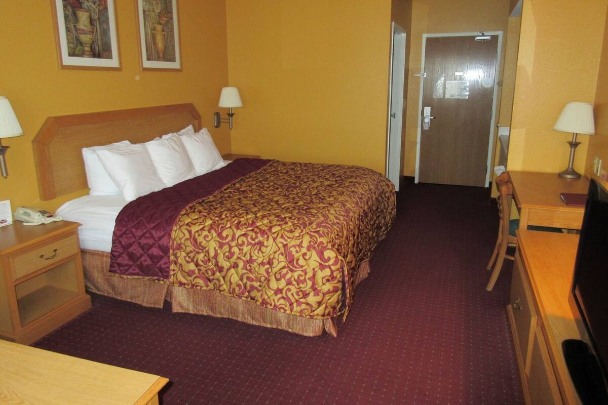 Habitación de hotel con cama, lámparas de noche, un escritorio con televisor y una puerta que da a otra habitación.