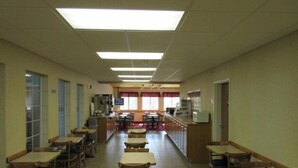 A spacious cafeteria with multiple tables and chairs, a counter with a display, and windows with red curtains.