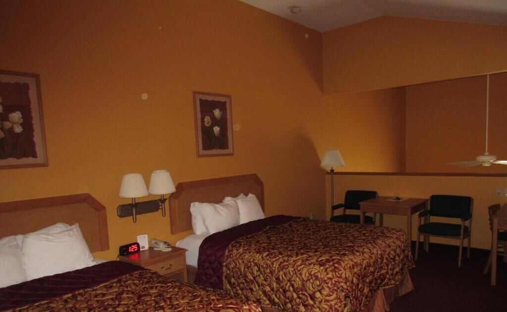 Habitación de hotel con dos camas, un escritorio y una silla. Hay lámparas de pared y un reloj sobre la mesita de noche.