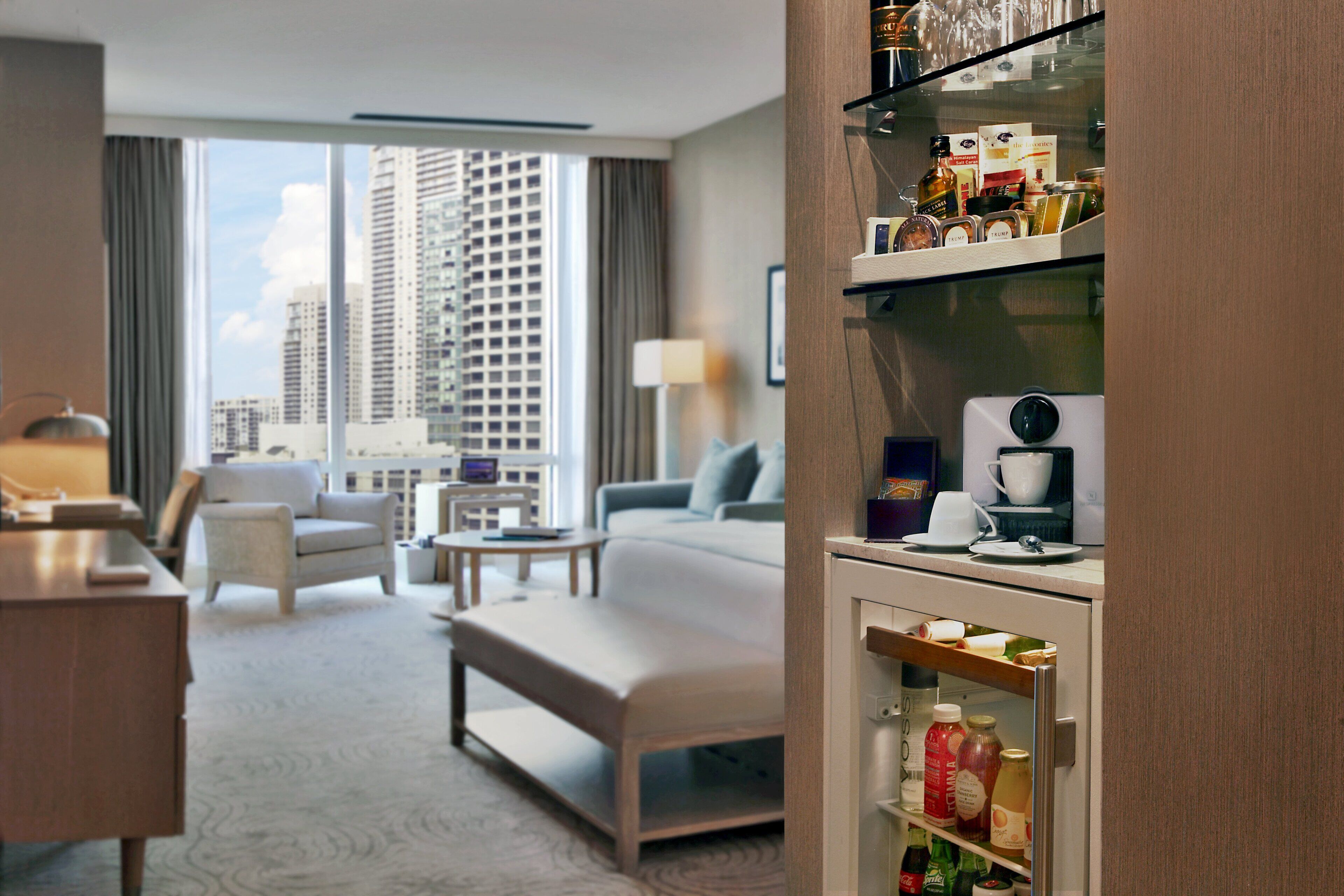 A hotel room with a large window, a bed, a desk, a chair, and a minibar.