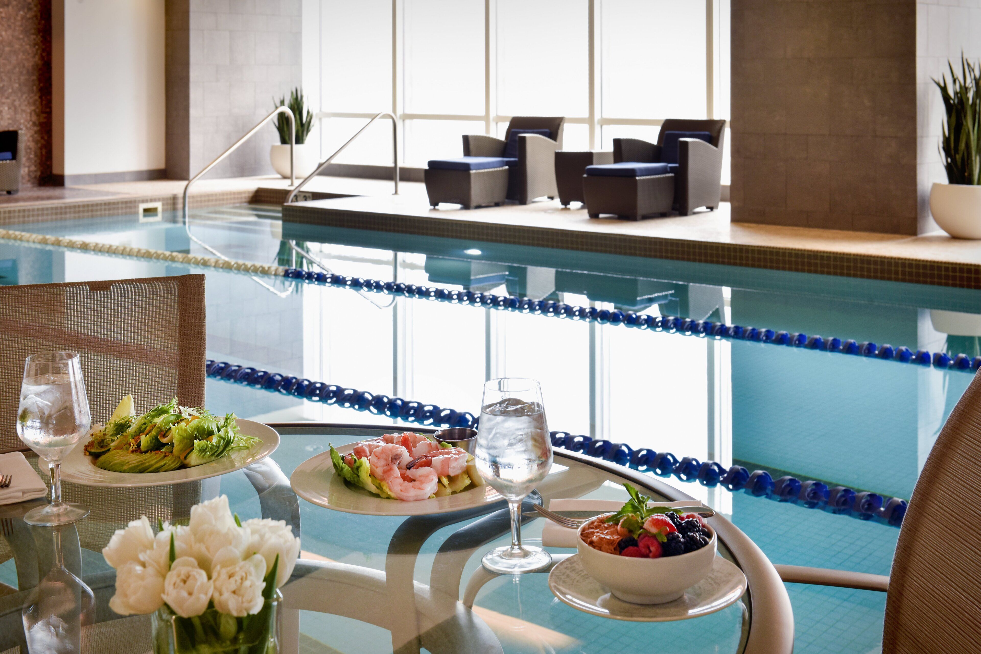 A poolside dining area with a table set for two, featuring a salad, shrimp, and a bowl of fruit.