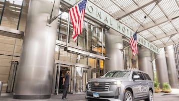 A modern hotel entrance with two American flags, a security guard, and a Cadillac SUV parked in front.