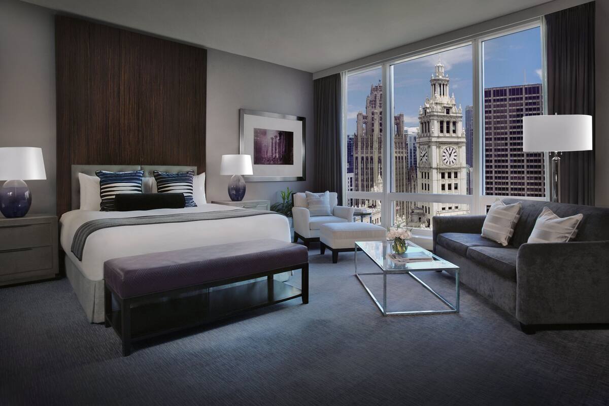 A modern hotel room with a large bed, a grey sofa, a coffee table, and a view of a cityscape with a clock tower.