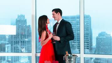 A couple in a red dress and a suit standing by a window with a city view.
