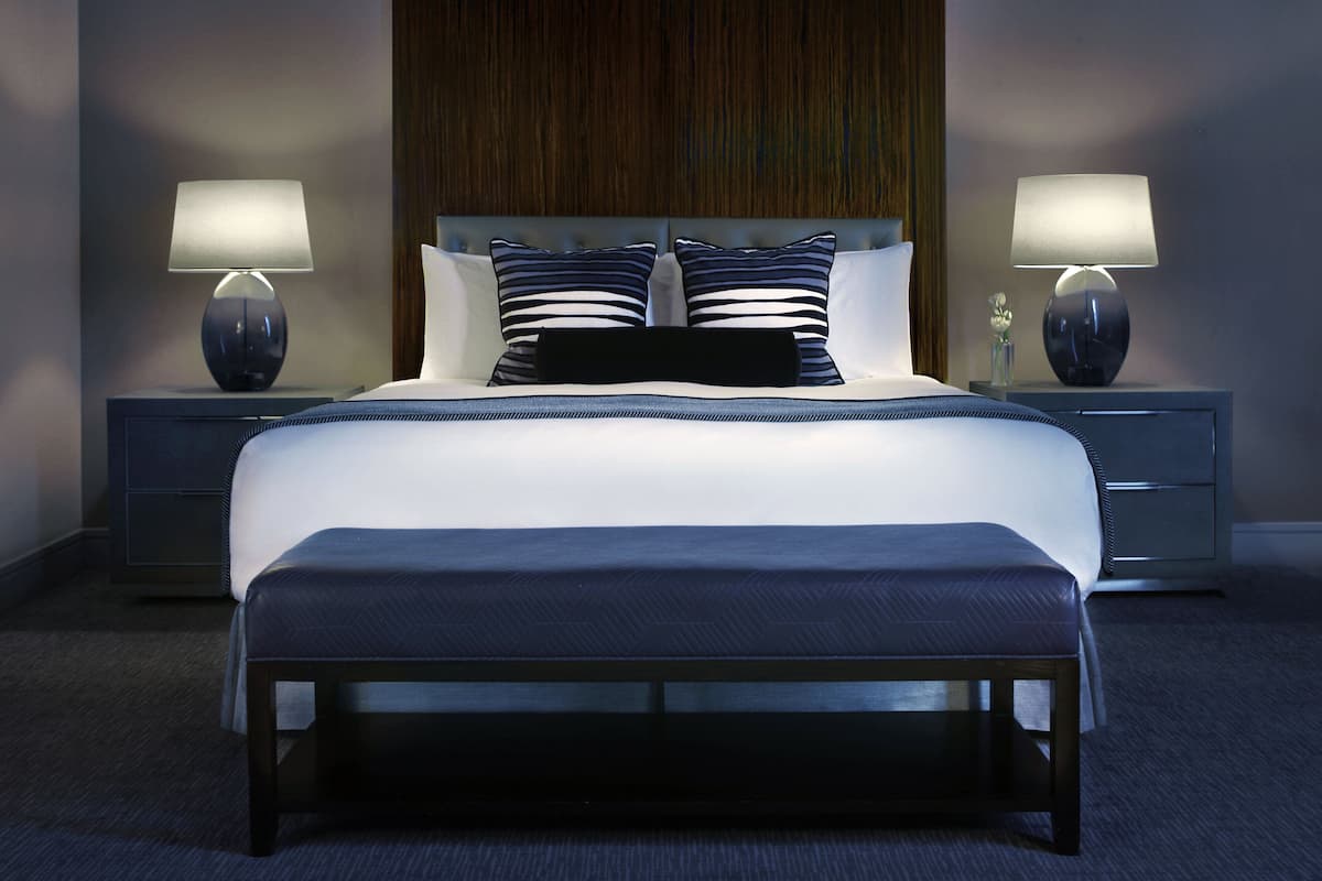 A modern hotel room with a large bed, two bedside tables with lamps, a wooden headboard, and a dark carpet.