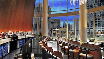 A modern bar with a curved counter, multiple seating areas, and large windows offering city views.