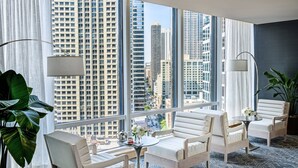 Una habitación de hotel moderna con ventanas amplias, mobiliario blanco y vistas a edificios altos.