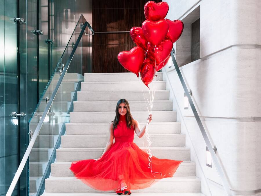 A person in a red dress sitting on a staircase with heart-shaped balloons.