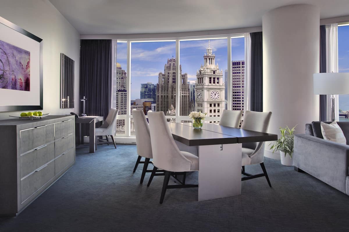 A modern hotel room with a dining area, large windows with city views, and a cityscape visible outside.