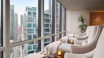 Una habitación de hotel moderna con un ventanal que ofrece vistas a la ciudad, una zona de estar acogedora con una planta y una pequeña mesa con una vela.