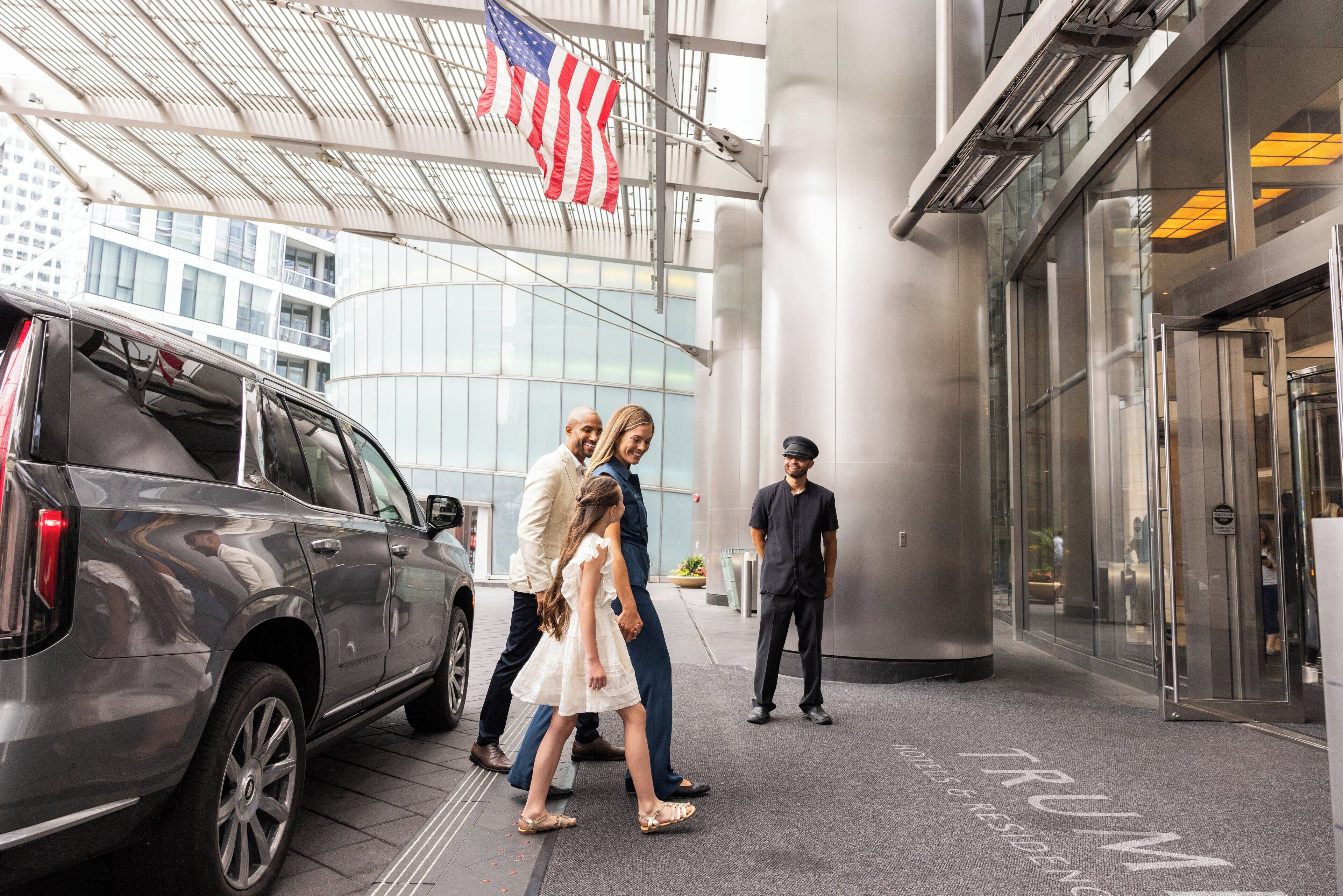 A hotel entrance with a gray SUV parked outside, a security guard, and a family walking towards the entrance.
