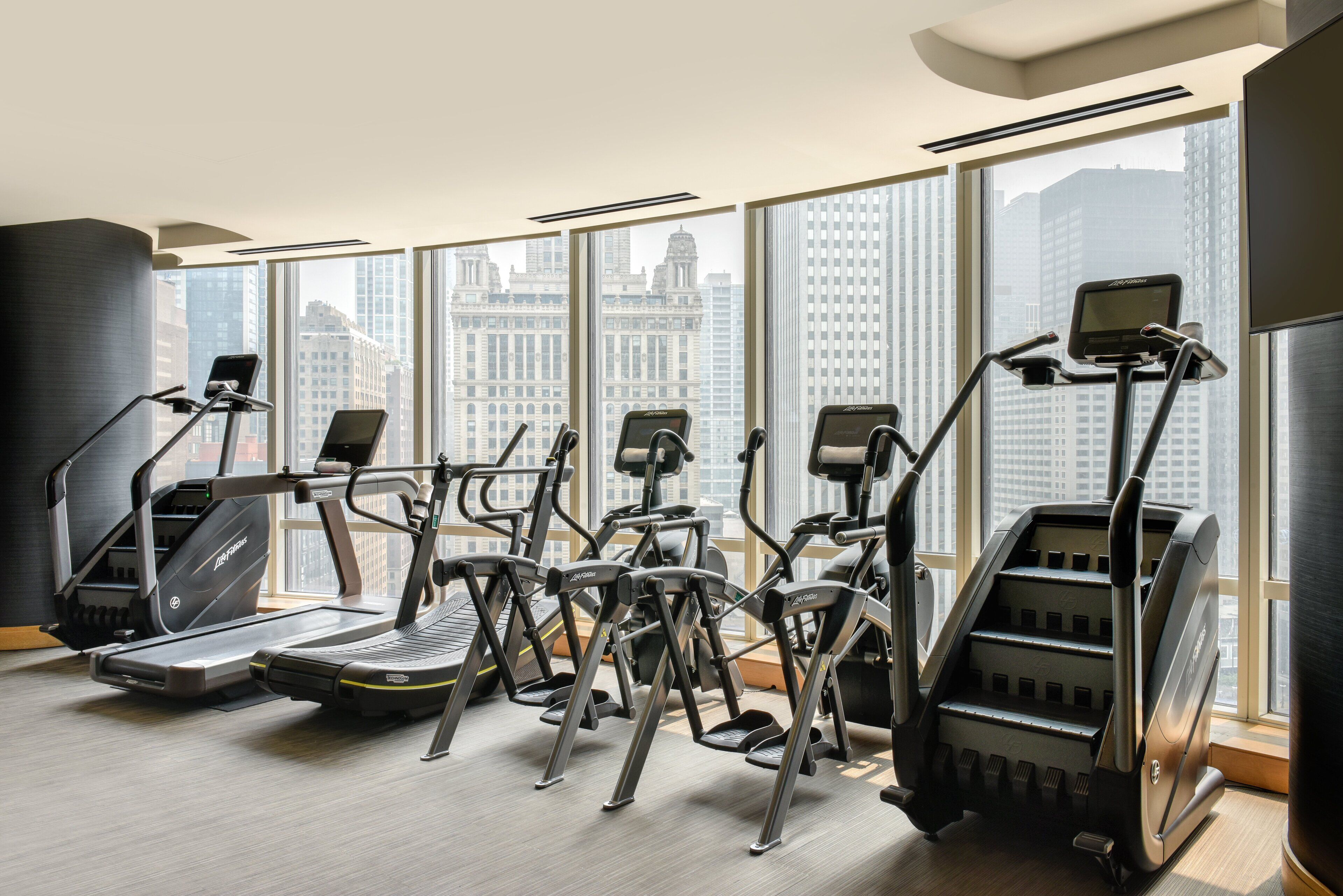 Ein Fitnessstudio mit Laufbändern, Crosstrainern und einer Steiganlage, große Fenster mit Blick auf die Stadt.