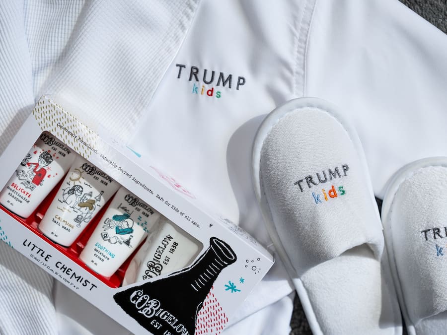 Un par de zapatillas blancas con el bordado "TRUMP kids", junto con una camisa blanca y una caja de productos para bebés.