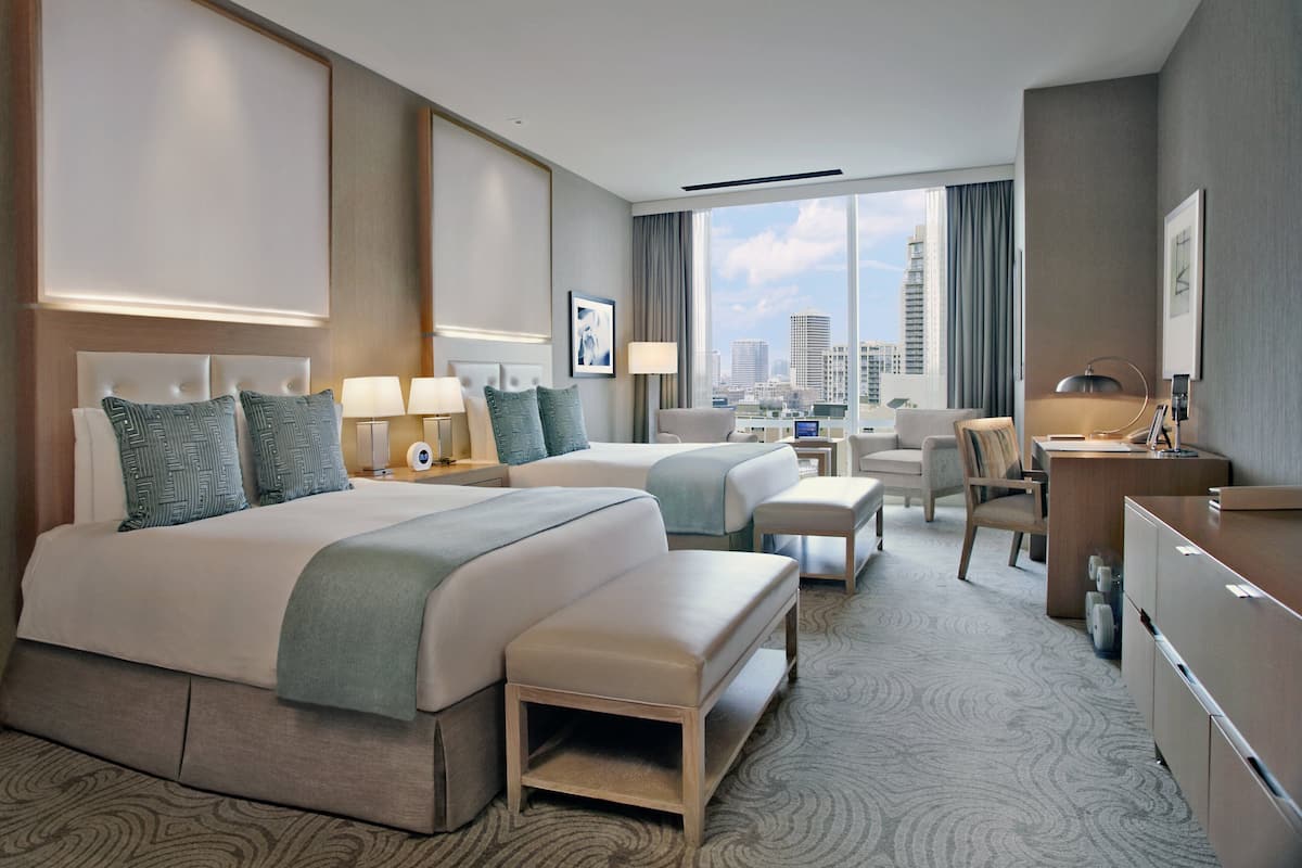 A hotel room with a large bed, two bedside tables with lamps, a desk, a chair, and a view of the city.