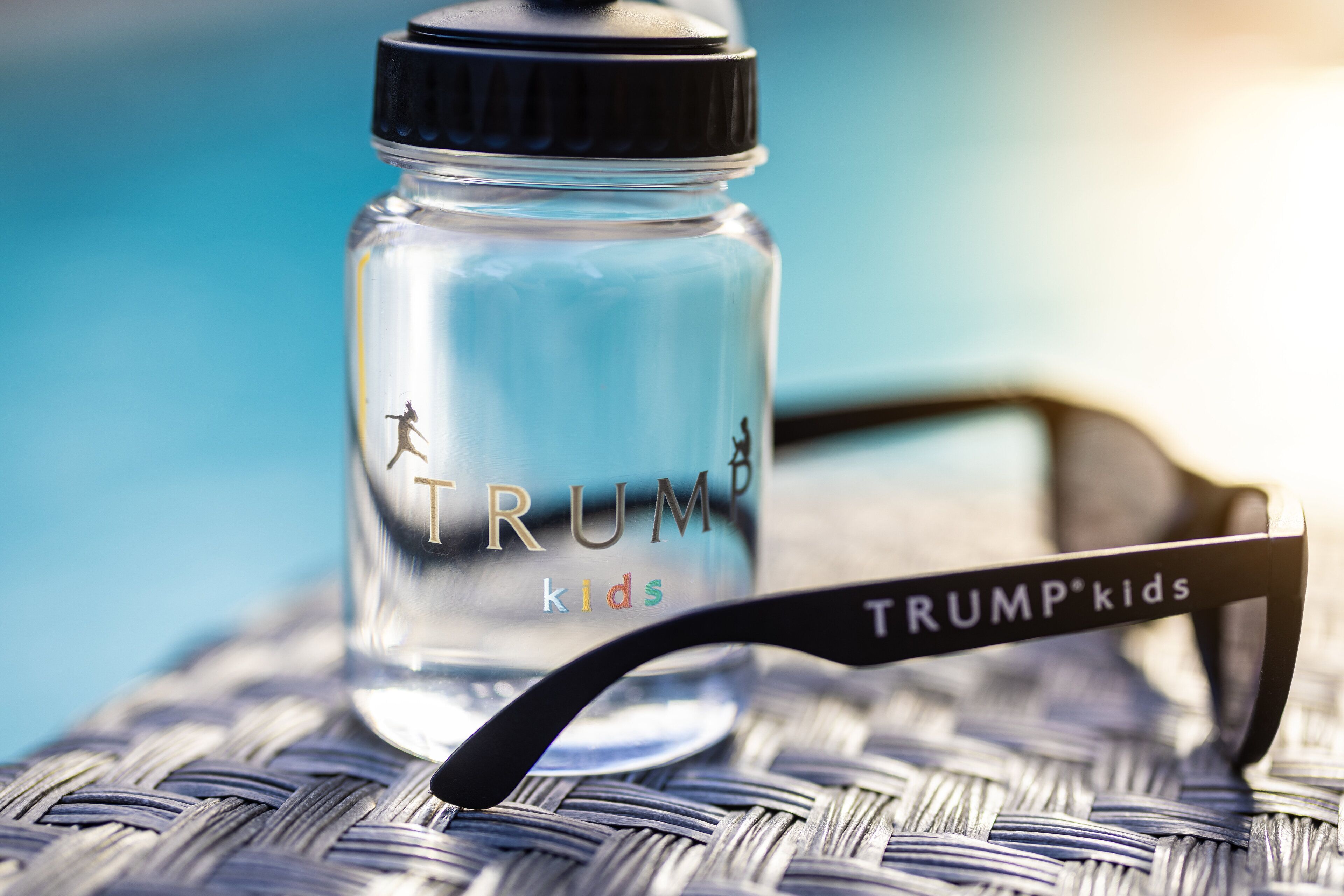 Une bouteille d’eau transparente en verre, avec un couvercle noir, et une paire de lunettes de soleil dont la monture porte l’inscription « TRUMP kids », le tout posé sur une surface tissée.