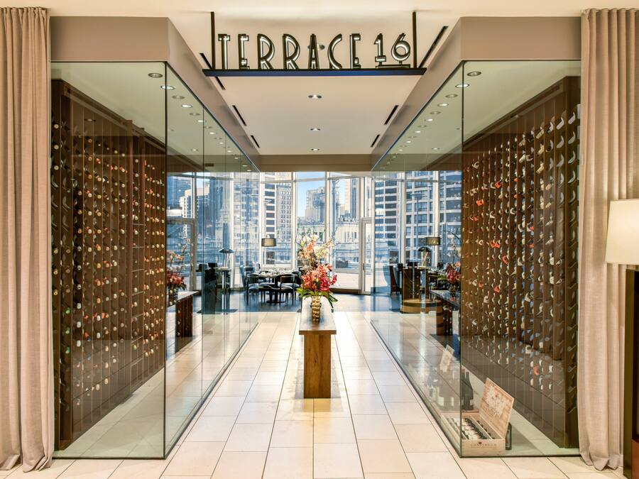 A modern hotel lobby with a glass wall displaying a wine rack, a central wooden table with flowers, and a view of the cityscape.