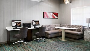 Una sala de reuniones moderna con un escritorio blanco, dos monitores de computadora, una silla de oficina gris, un sofá estampado y una alfombra con diseño.
