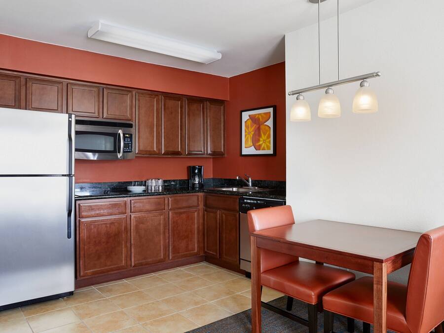A compact kitchen with wooden cabinets, a stainless steel refrigerator, a microwave, and a dishwasher. There is a dining table with two chairs.
