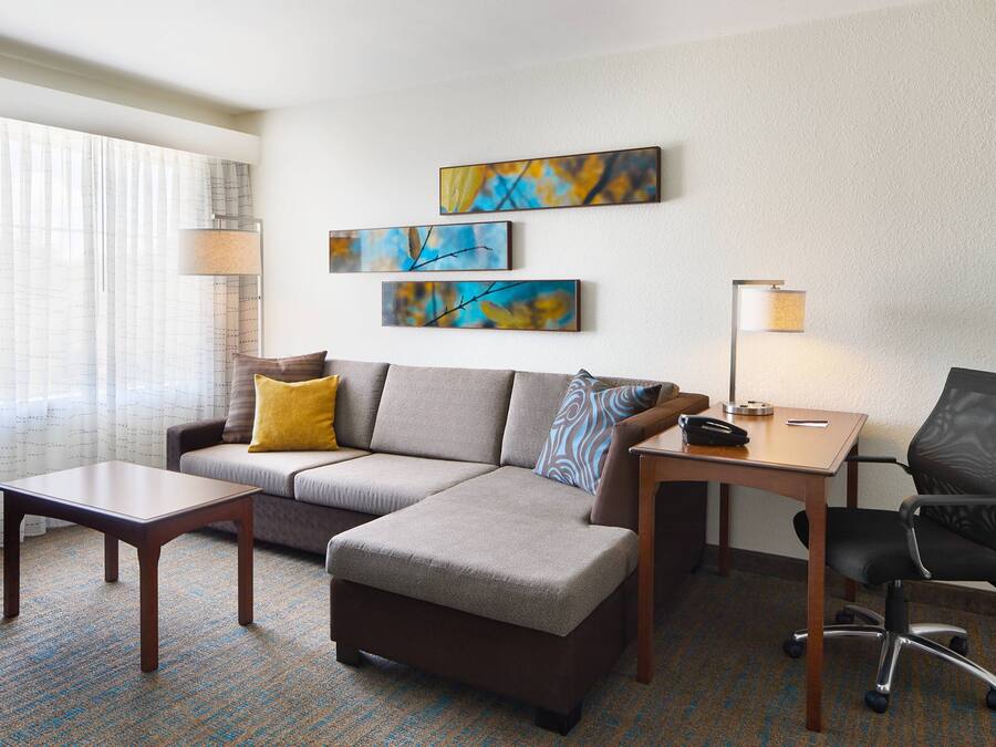 A modern hotel room with a sectional sofa, a small table, a desk, and a chair. There are three abstract paintings on the wall and a window with curtains.