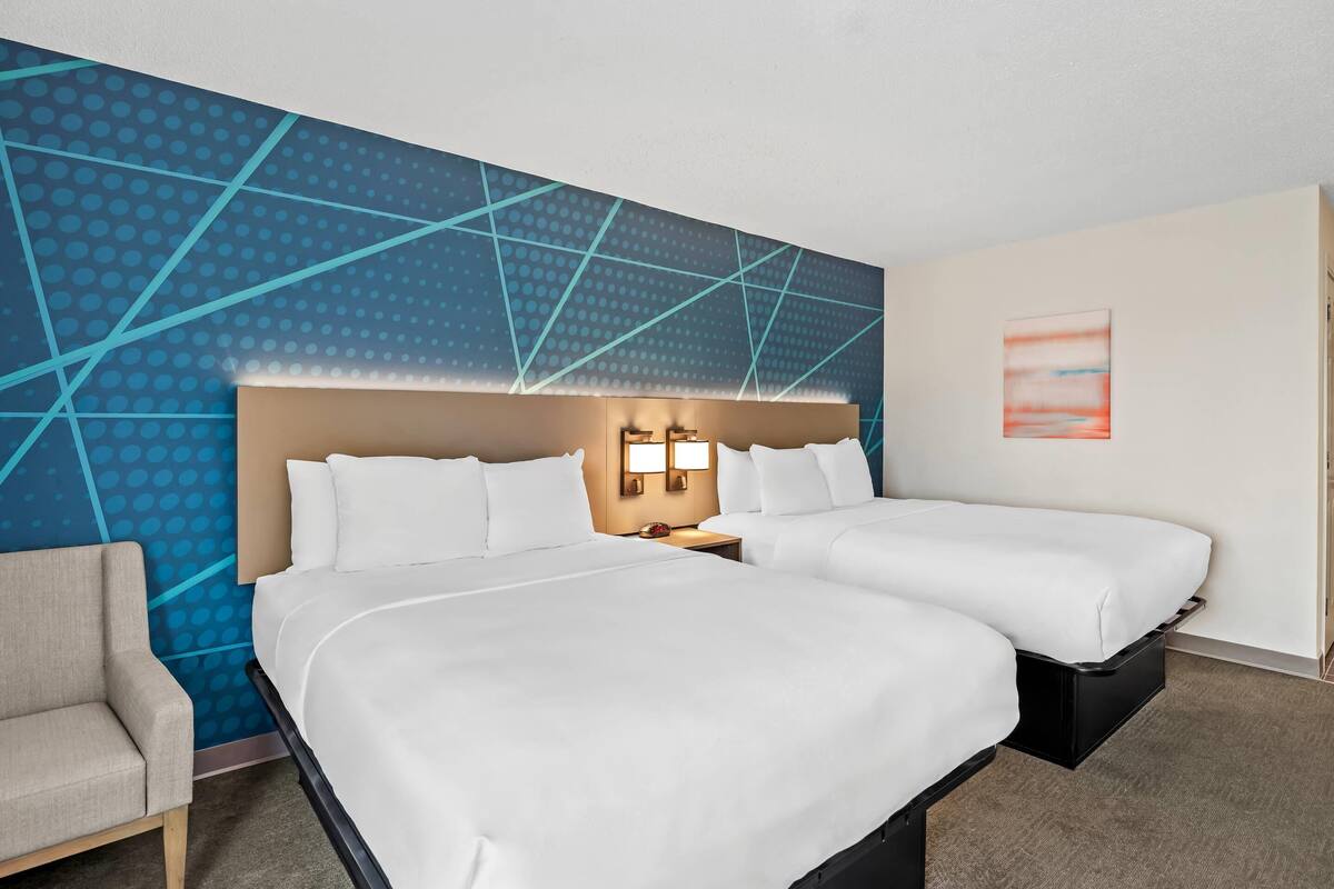 A hotel room with two beds, a nightstand, a lamp, a chair, and a blue patterned wall.