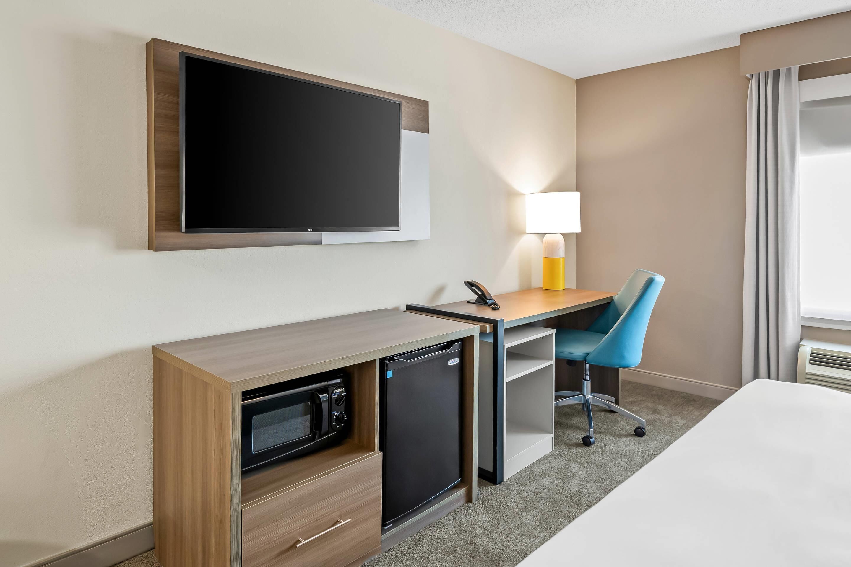 A hotel room with a flat-screen TV mounted on the wall, a desk with a lamp, a microwave, a mini fridge, a blue chair, and a bed.