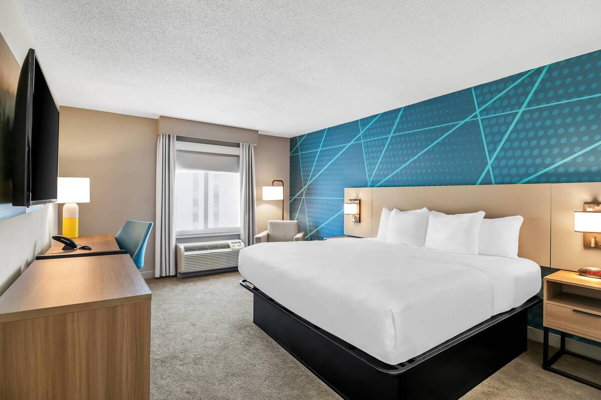 A hotel room with a bed, desk, chair, TV, and a wall with a geometric pattern.
