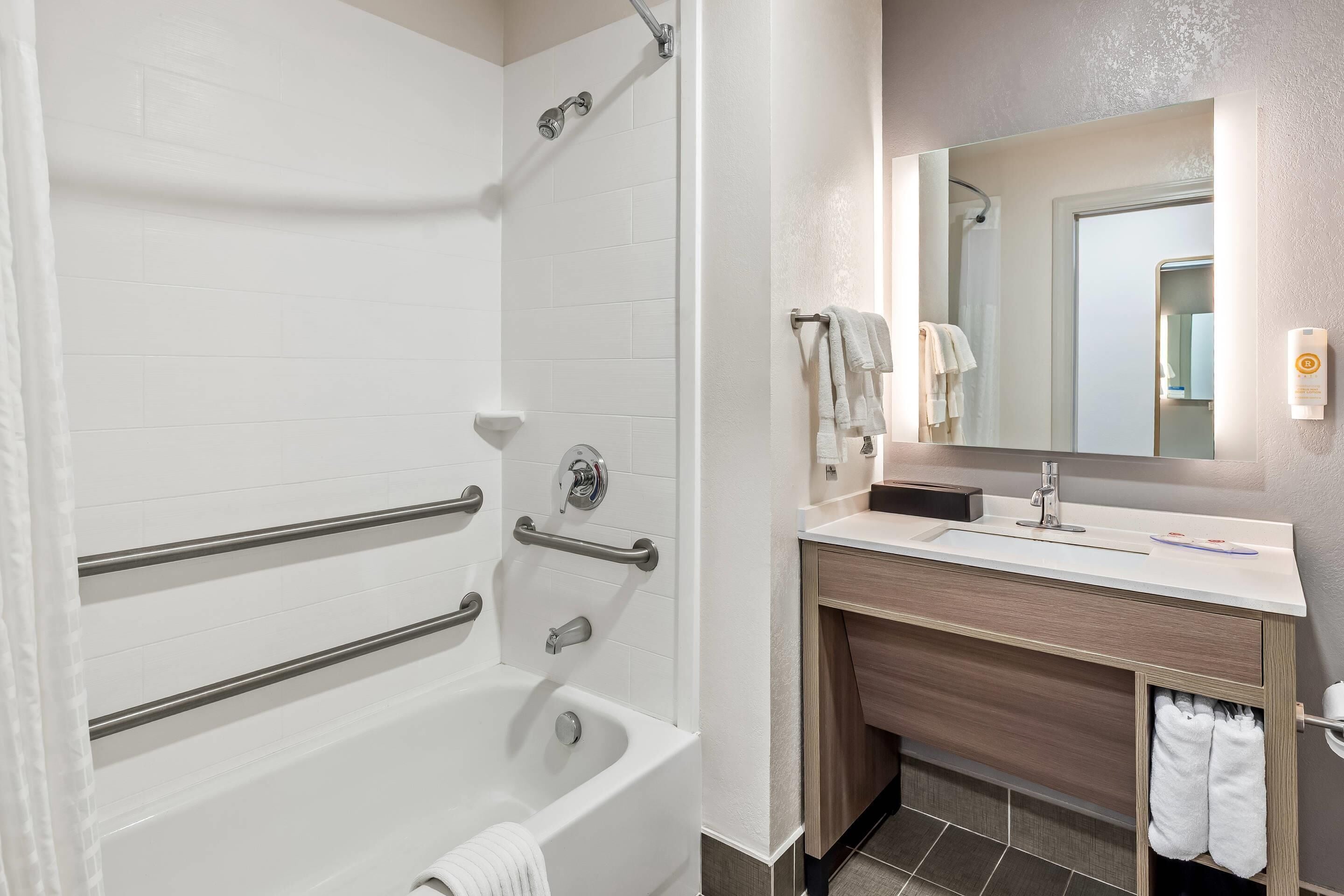 Un baño moderno con bañera, barras de apoyo, lavabo con espejo y un perchero para toallas.